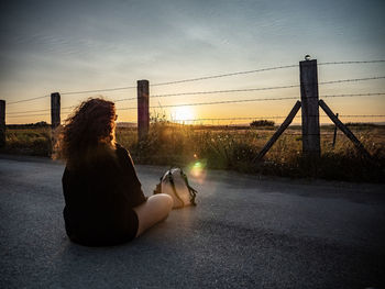 Rear view of woman sitting on road against sky during sunset
