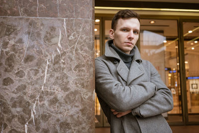 A young man stands outside a shop on the street in a gray coat leaning.