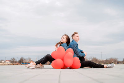 Young couple sitting back to back in the street holding a pile of red balloons spending time