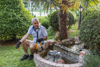 Garden. a mature man by the small fountain has a break while working in the tools.