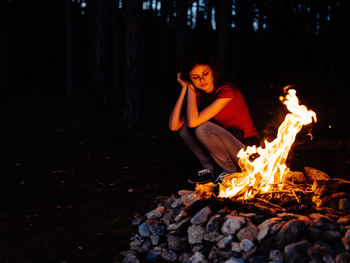 Young woman sitting by bonfire in forest at night