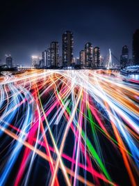 Light trails on street by illuminated buildings against sky at night