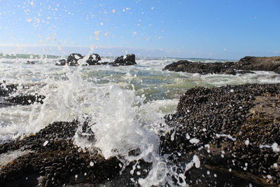 Waves splashing on rocks at shore against clear sky