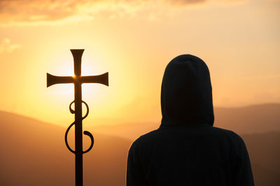 Dark silhouette of person in a hood and christian cross against sun on the background