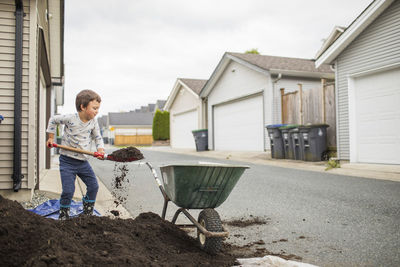 Young boy lifting shovel full of soil into wheelbarrow in back alley