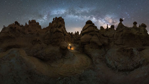 Silhouette of unrecognizable explorer standing with flashlight on scenery of rocky formations in highlands under milky way starry sky in goblin valley state park, utah, usa.