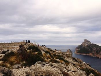 People on cliff by sea against cloudy sky