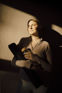 Woman with eyes closed standing against wall with sunlight on face