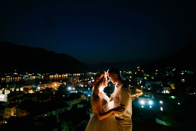 Woman standing by illuminated cityscape against sky at night