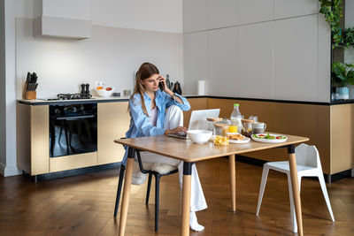 Busy woman freelancer has smartphone conversation working on laptop on kitchen at home.