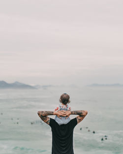 Rear view of man standing in sea against sky with his daughter