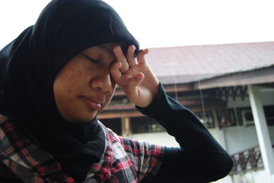 Close-up of young woman rubbing eyes while standing against house