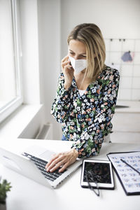 Businesswoman wearing mask using laptop while talking on phone at home