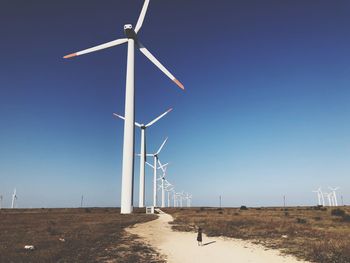 Girl walking on sand with windmill on field against clear blue sky