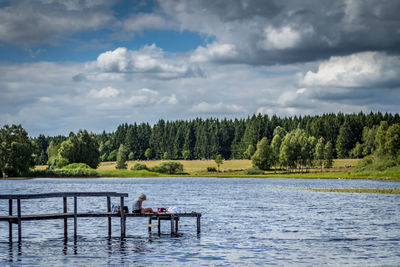 Woman sitting on pier over lake by trees against cloudy sky