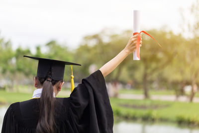 Rear view of young woman with hand raised holding certificate while standing in park