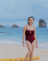 Smiling young woman wearing swimwear while standing at beach against cloudy sky