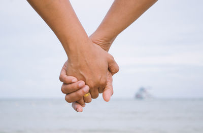 Cropped image of couple holding hands at beach against sky