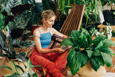 A young woman is sitting surrounded by potted plants on the floor and shopping on a digital tablet.