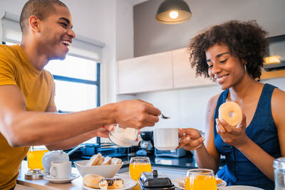 Cheerful man putting sugar in young woman coffee cup in kitchen