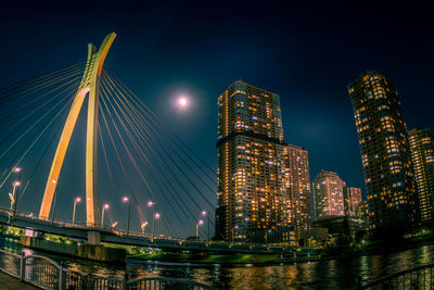 Illuminated modern buildings by river against sky at night