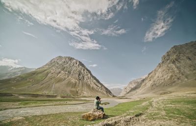 Side view of man photographing mountain while sitting on rock against sky