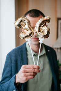 Man in a blue jacket holds in his hands little foiled gold balloons of the number 36 on sticks