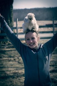 Portrait of smiling girl with silkie chicken on head standing at field