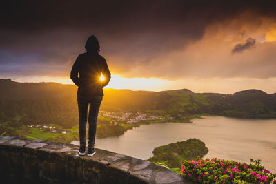 Rear view of woman standing on stone wall against sky during sunset