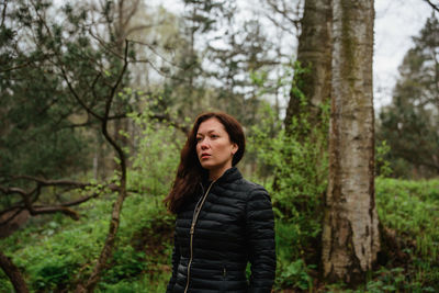 Portrait of young woman standing by tree trunk in forest