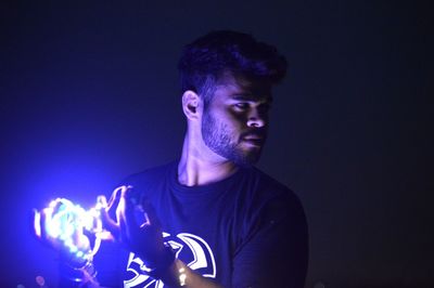 Young man holding illuminated lighting equipment while standing in darkroom