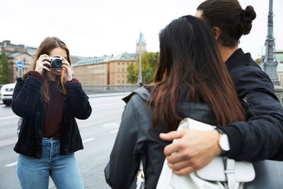 Woman photographing male and female friends while standing by street in city