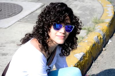 Young woman with curly hair wearing sunglasses while sitting on footpath