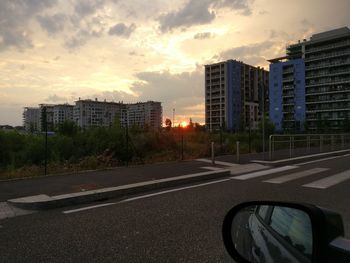 Road by buildings against sky during sunset