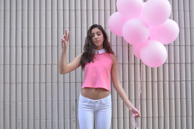 Woman holding pink helium balloons while gesturing peace sign by patterned wall