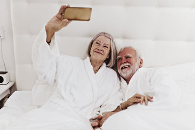 Smiling senior woman taking selfie through mobile phone with man while lying on bed in hotel room