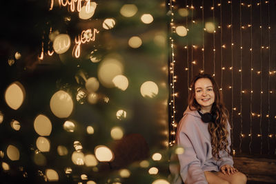 Young smiling woman with headphones, sitting near glowing christmas tree, bokeh foreground, evening