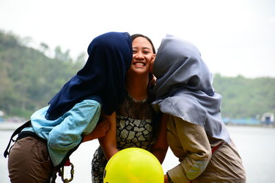 Women kissing smiling friend standing against lake