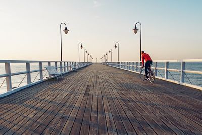 Rear view of man cycling on pier at sea against clear sky