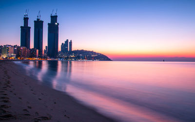 Sea by illuminated buildings against sky during sunset