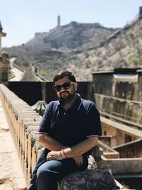 Portrait of young man sitting on retaining wall against sky