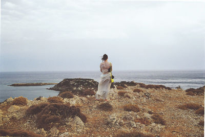 Rear view of woman looking at sea while standing on rock against sky