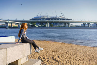 Beautiful redhead young woman sits on the embankment on a spring day