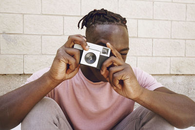Man photographing through camera while sitting against wall