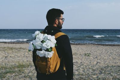 Man standing on beach with flowers on backpack against clear sky