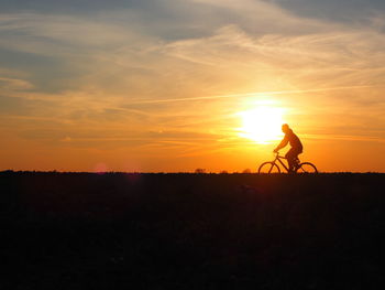 Silhouette person riding bicycle on field against sky during sunset