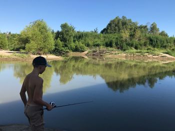 Full length of shirtless man fishing in lake against sky