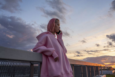 Young woman with pink hair wearing pink hooded shirt leaning against railing and talking on mobile phone