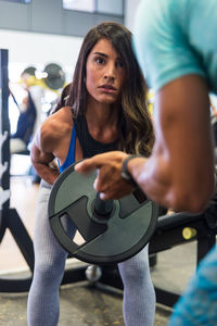 Concentrated young ethnic female in sportswear doing exercises with barbell with support of personal instructor during training in gym