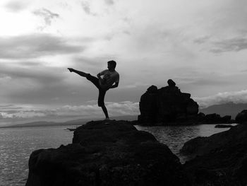 Silhouette young man standing on rock by sea against sky